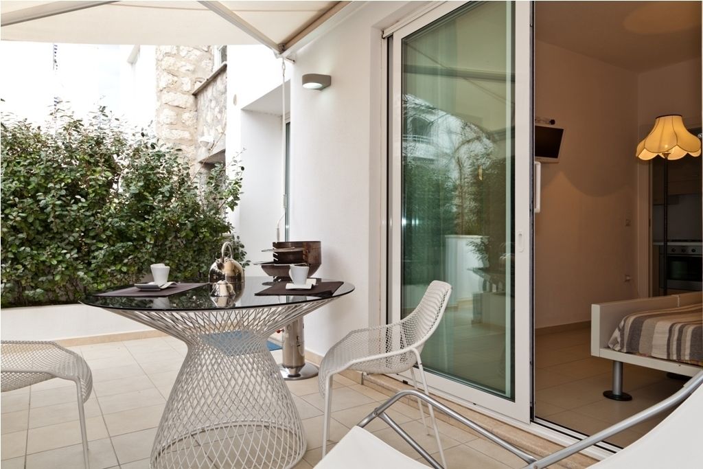 A modern outdoor patio with a glass table, white chairs, and a glass door leading to an interior space.