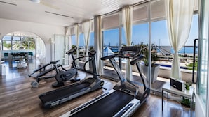 Un gimnasio con cintas de correr, bicicletas estáticas y pesas, ventanas amplias con cortinas y vistas al mar.