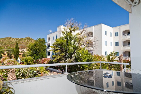 A balcony with a glass table, surrounded by lush greenery and white buildings with blue shutters.