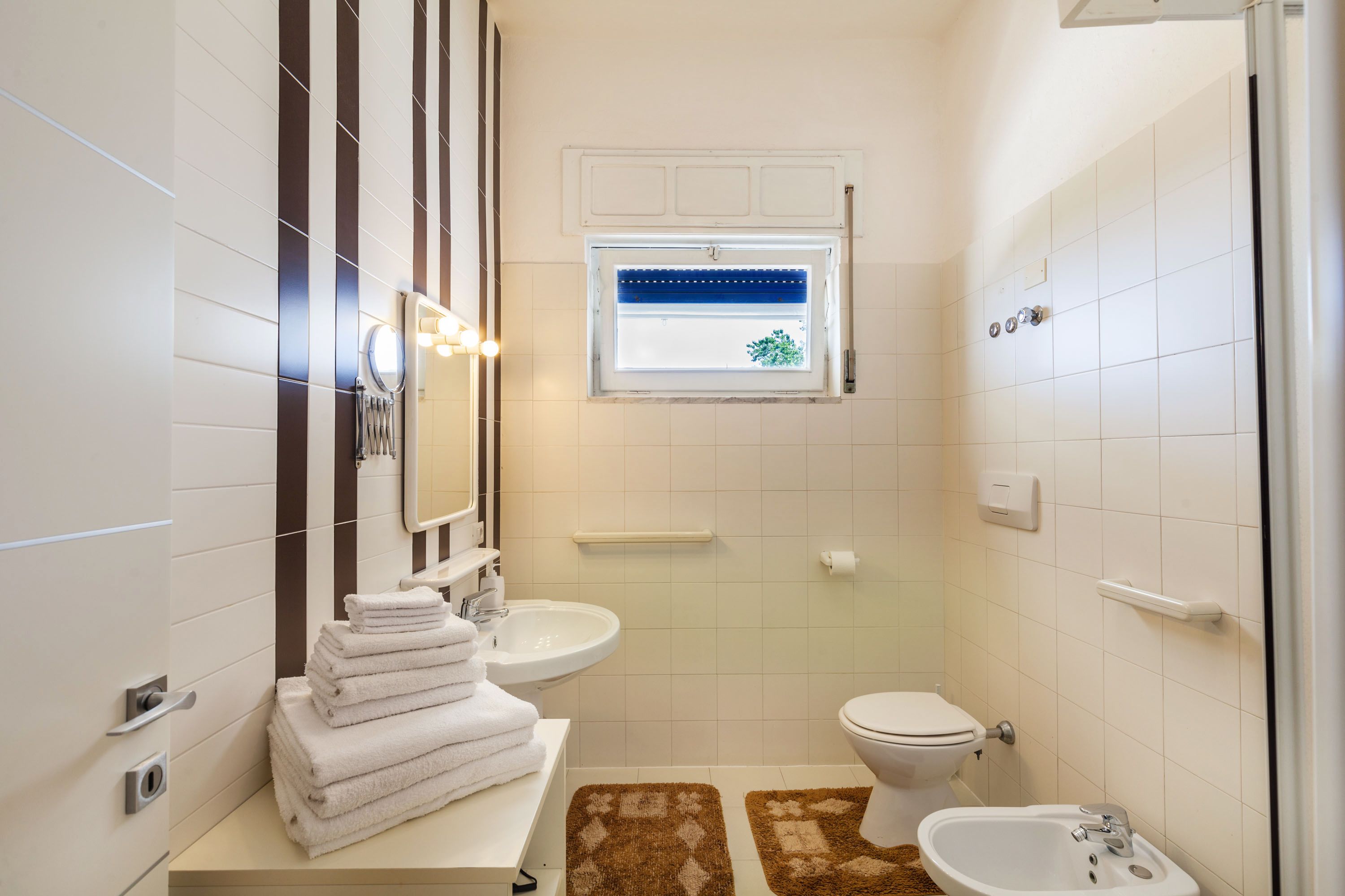 Un baño con inodoro, lavamanos y espejo. Hay toallas apiladas en el mostrador y una ventana sobre el espejo.