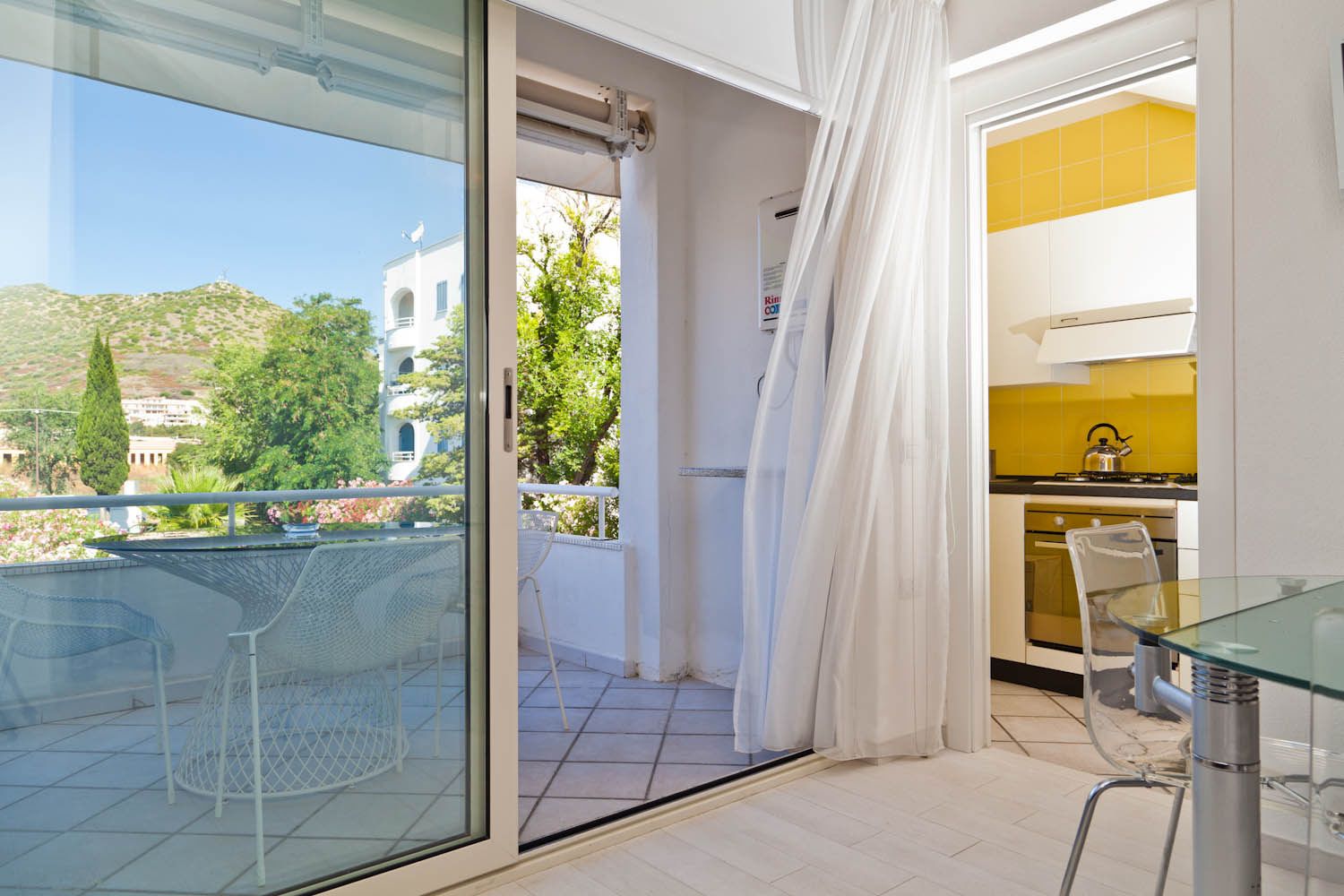 Ein Balkon mit Glastisch und Stühlen, Blick auf eine Hügellandschaft und eine Küche mit gelben Fliesen.