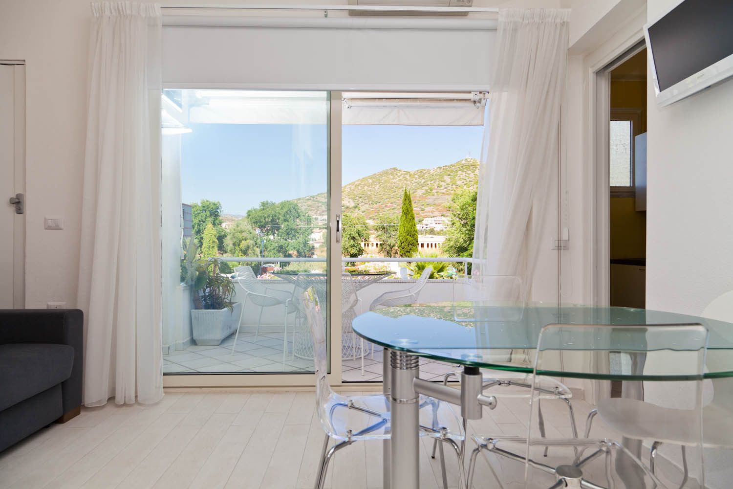 A modern living room with a glass table and chairs, a grey sofa, and a sliding glass door leading to a balcony with a view of a hillside and trees.