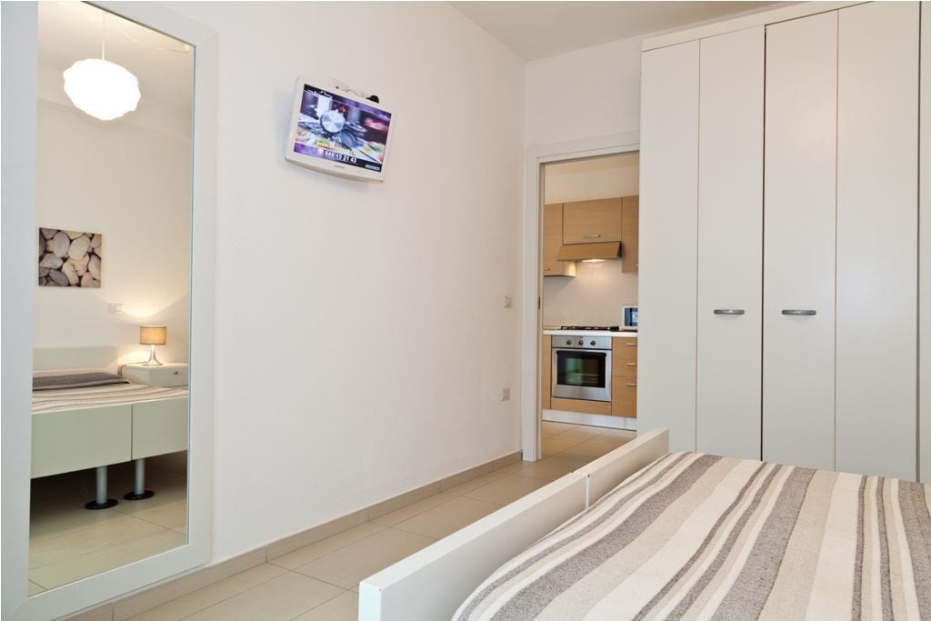 A bedroom with a bed, a bedside table, a lamp, a TV mounted on the wall, and a view into the kitchen area.