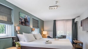 A hotel room with a large bed, bedside lamps, a small table, a chair, and a window with curtains. - Paviljoen Hotel (Rhenen)