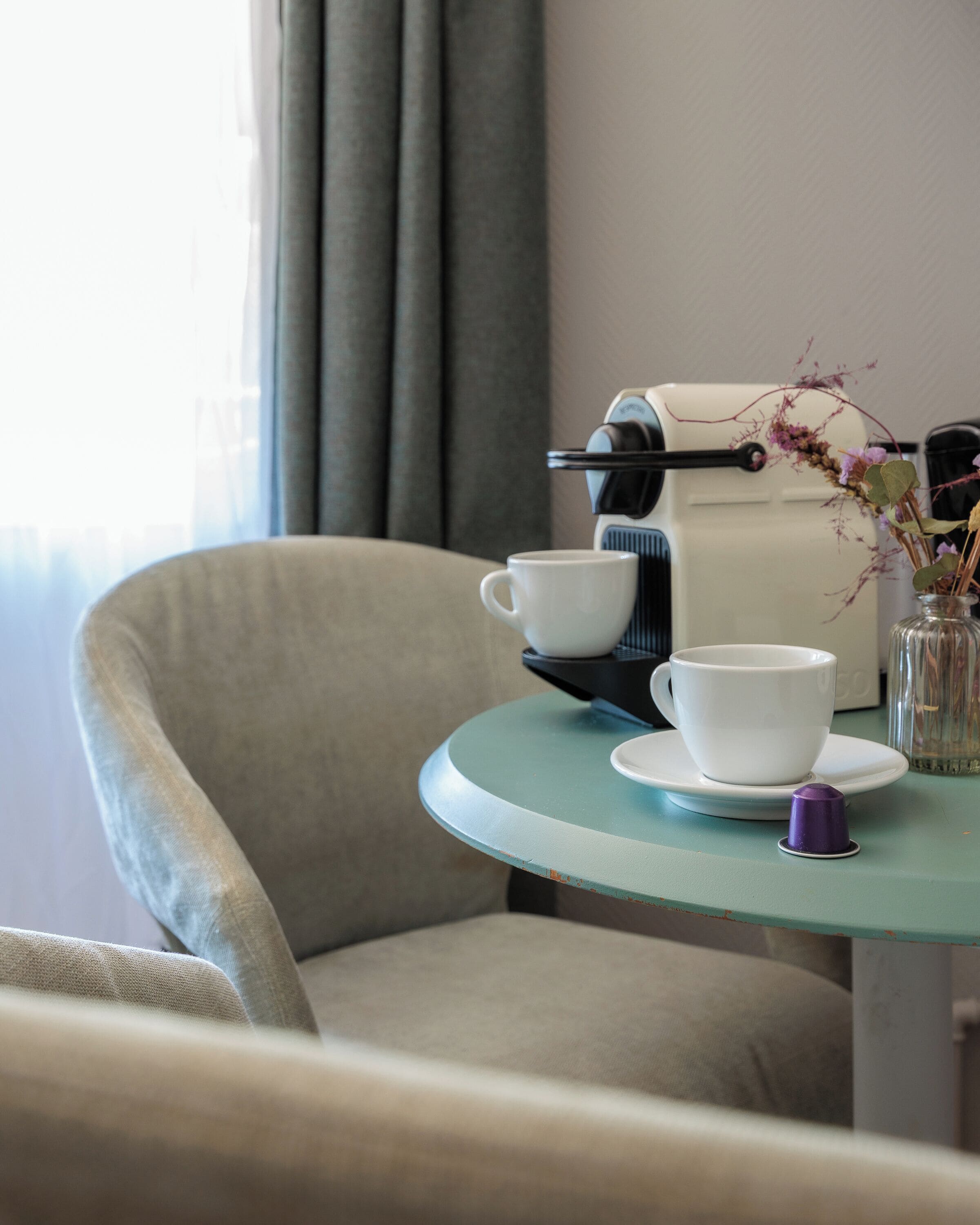 A room with a table set for two, a coffee machine, a cup, a saucer, and a vase with flowers.