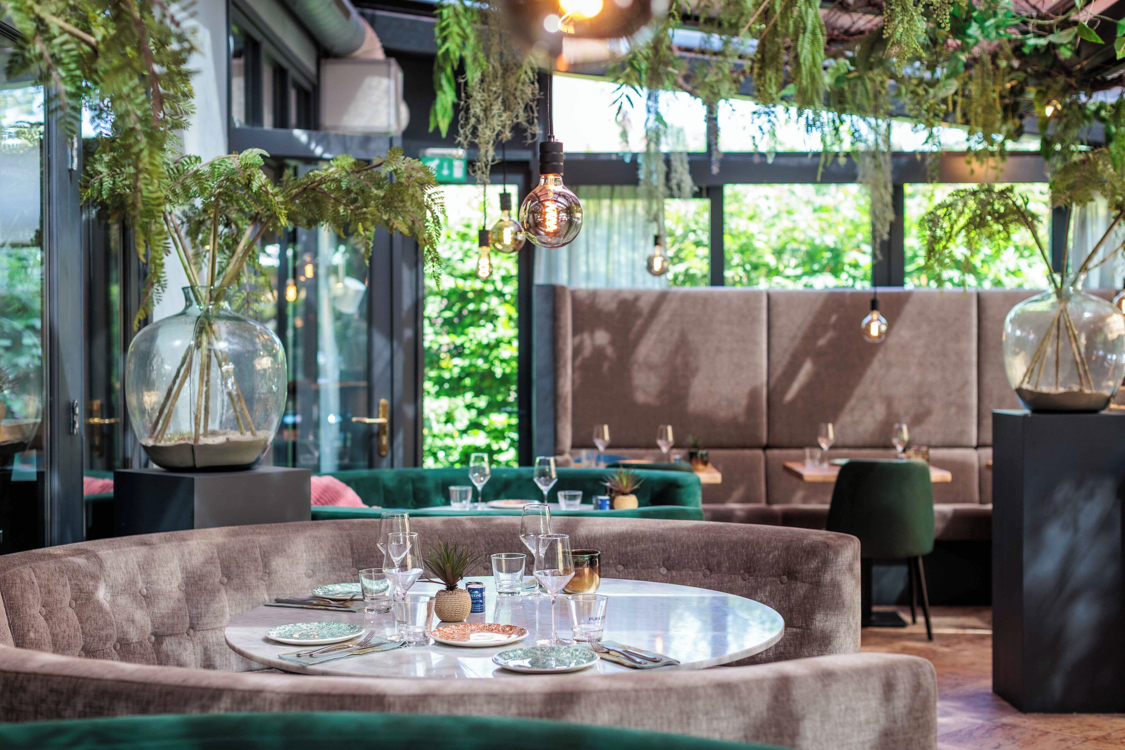 Een eetruimte met ronde tafels, beklede zitplaatsen in groen en hangplanten.