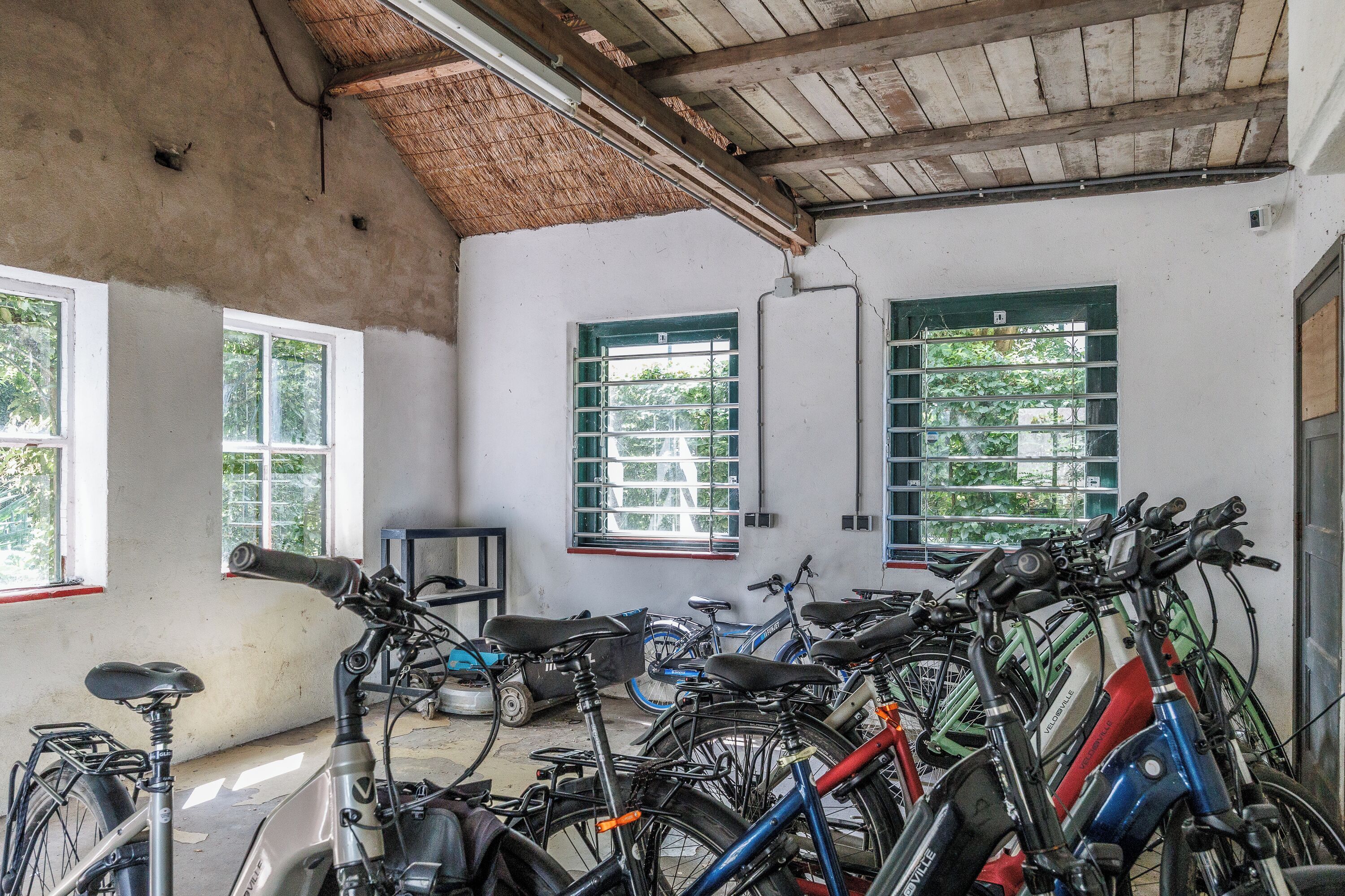 A room with a concrete ceiling, white walls, and several windows with green shutters. Multiple bicycles are parked in the room.