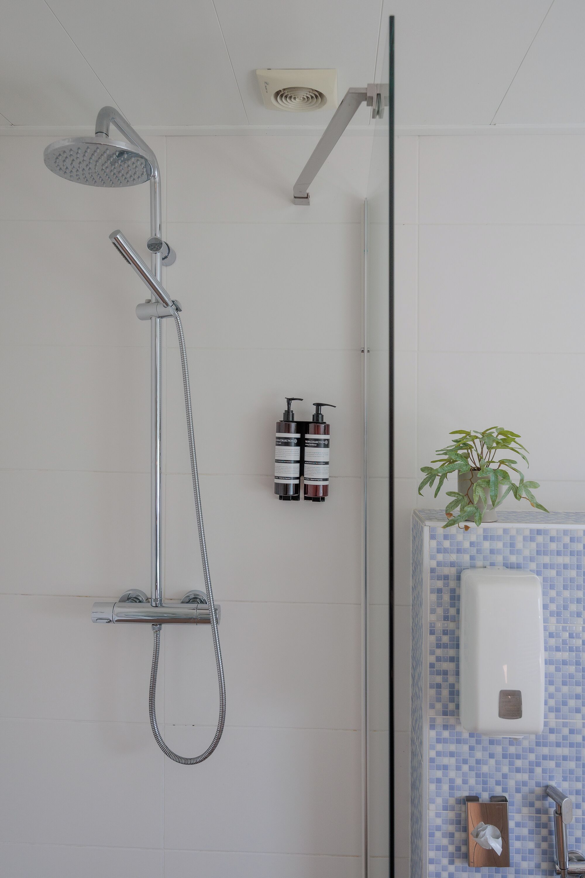 A shower with a handheld showerhead and a wall-mounted faucet, a hand dryer, and a soap dispenser on a tiled wall.