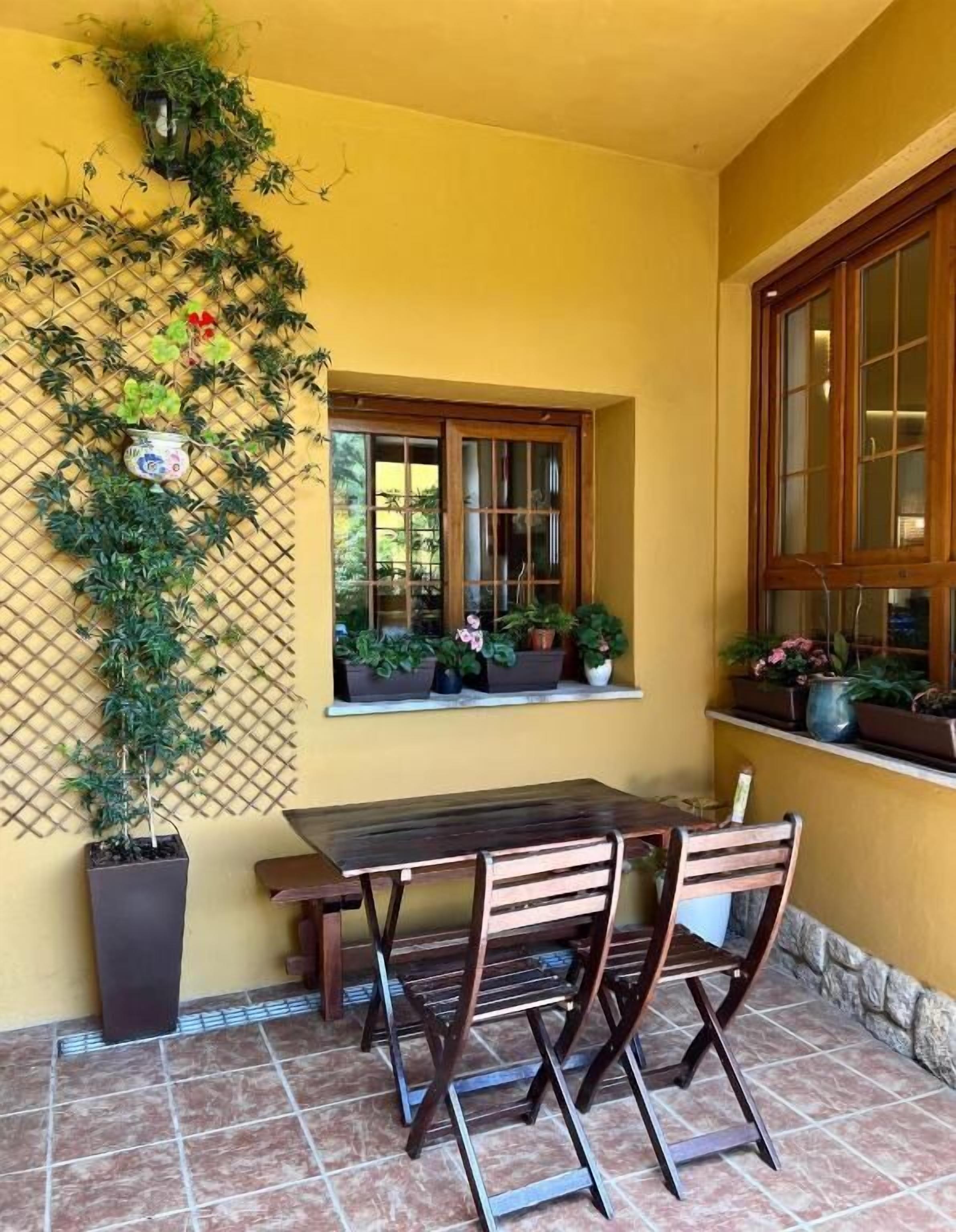 Une terrasse avec une table et des chaises en bois, une fenêtre ornée de plantes en pot et un mur en treillis recouvert de plantes grimpantes.