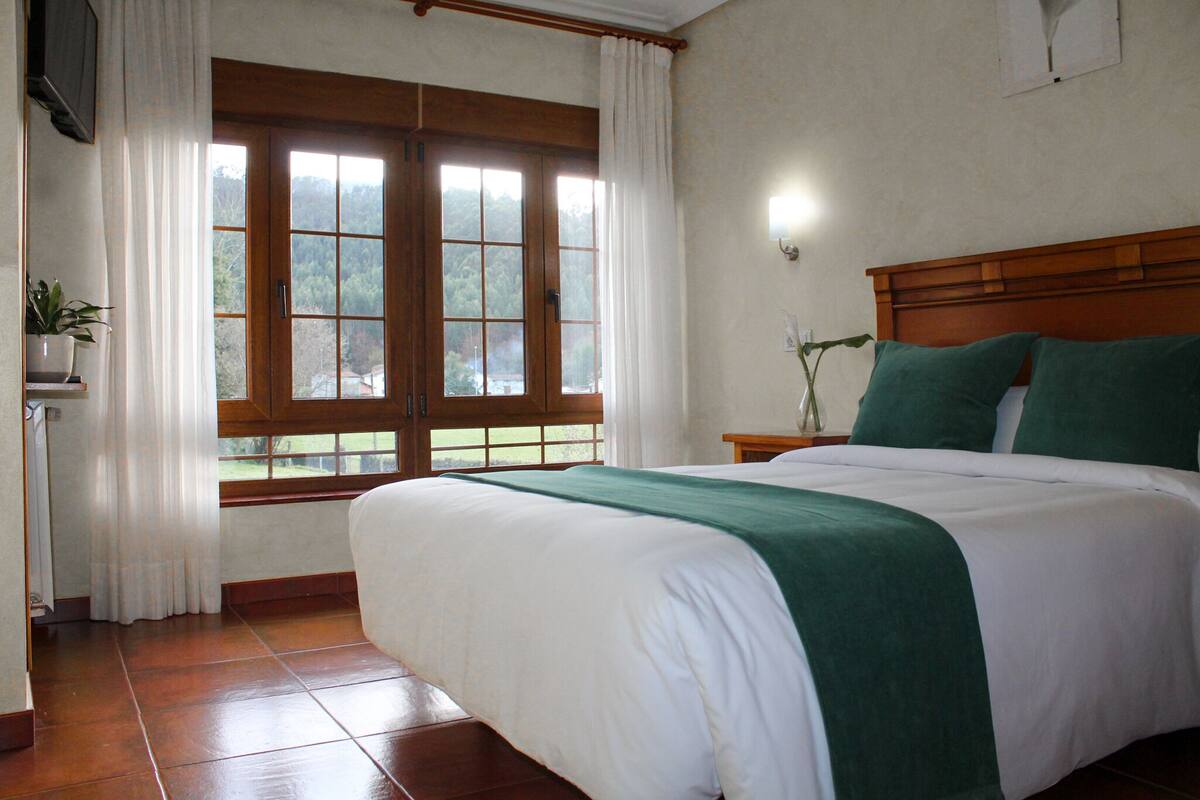 A bedroom with a large bed, a wooden headboard, a window with curtains, and a bedside table with a plant.