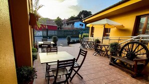 An outdoor patio with wooden tables and chairs, a yellow building in the background, and a large wheel structure. - Hotel Pugide (Llanes)