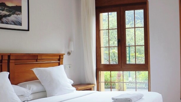 A neatly made bed with white linens and towels, a wooden headboard, a nightstand with a lamp, and a large window with a view of greenery. - Hotel Pugide (Llanes)