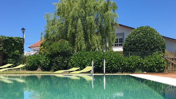 A clear swimming pool with tiled edges, surrounded by lush greenery and a white building with a red roof. - La Regalina (Valdes)