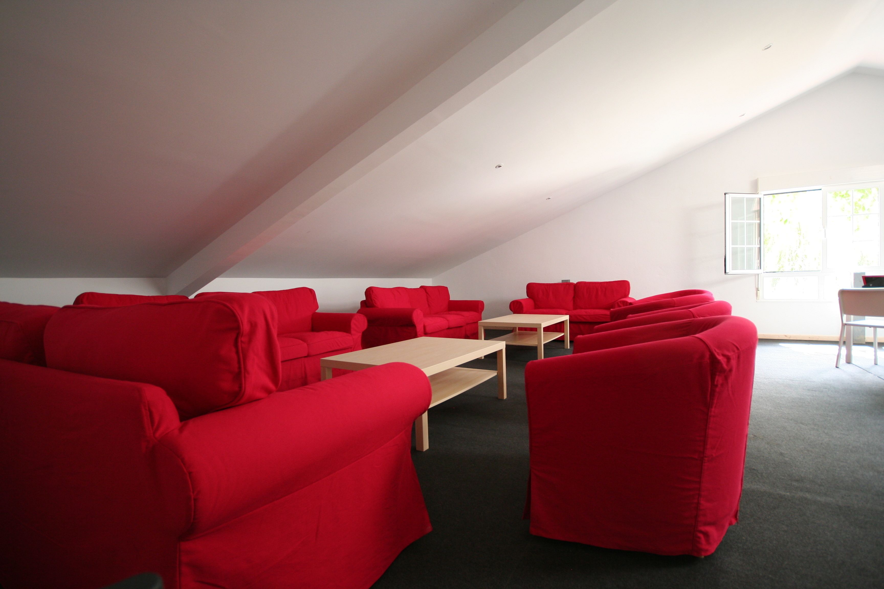 Une pièce dont le mobilier, notamment les canapés et la table, est recouvert d’un tissu rouge. La pièce possède un plafond en pente et une fenêtre qui laisse entrer la lumière naturelle.