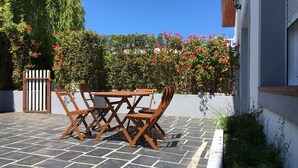 A patio with a wooden table and chairs, surrounded by a white wall and a hedge with red flowers. - La Regalina (Valdes)