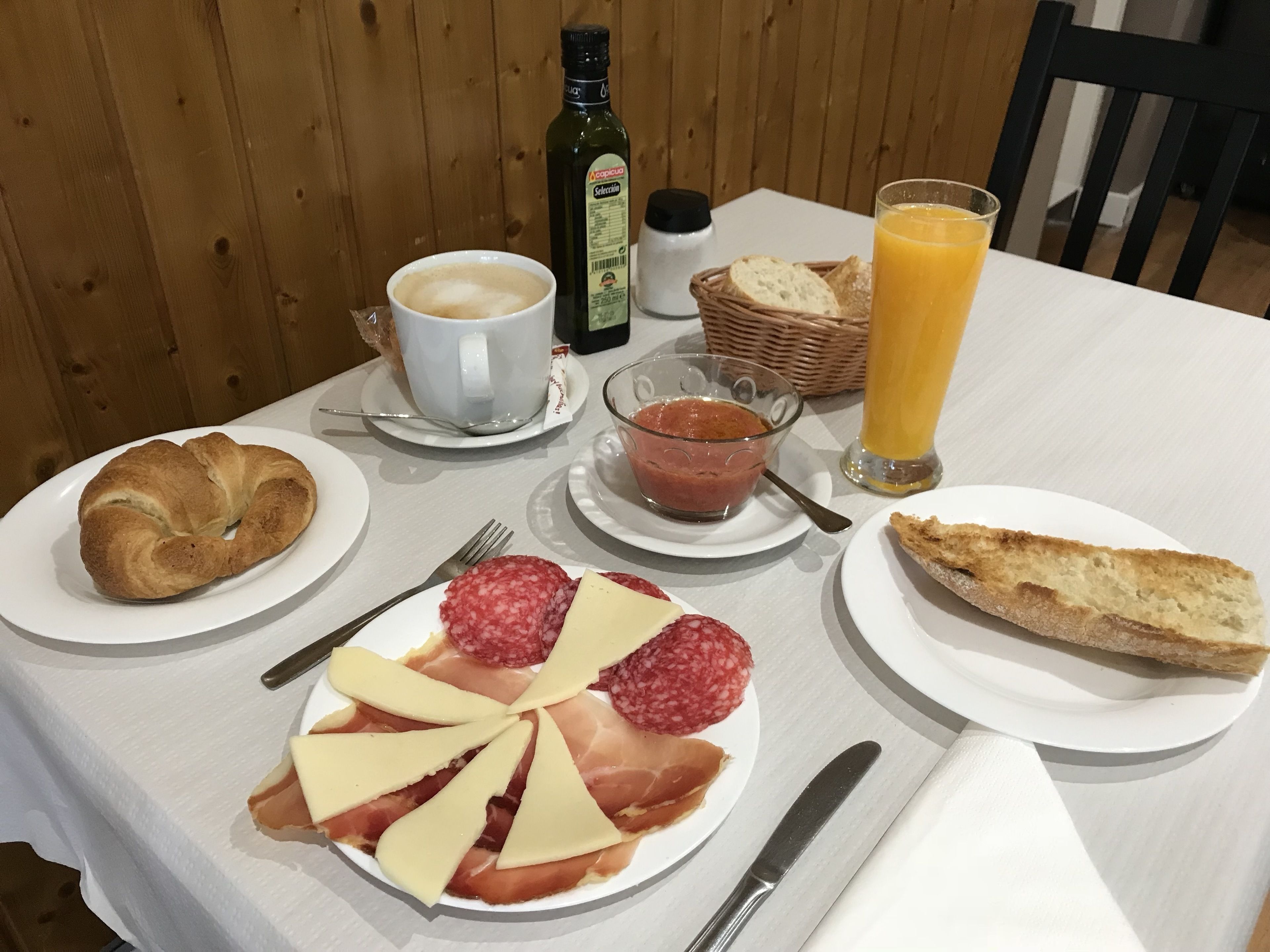 Une table de petit-déjeuner avec des croissants, du fromage, du salami et un verre de jus d’orange.