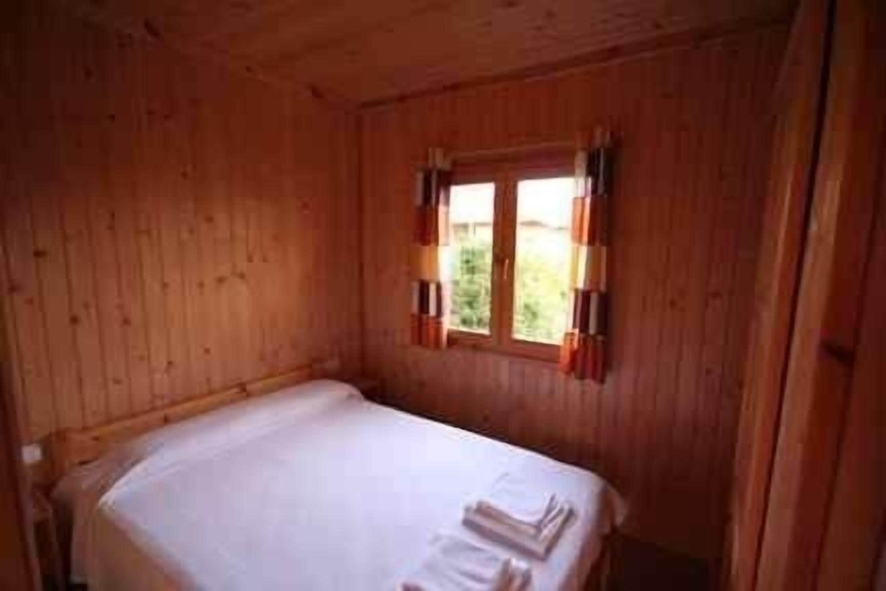 A wooden room with a single bed, a window with curtains, and a small table.
