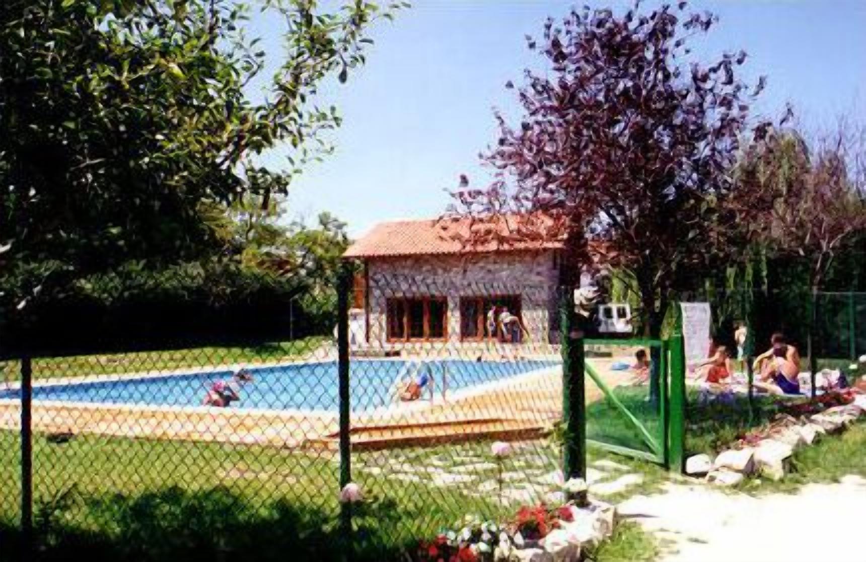 Un área de piscina con un deck cercado, un pequeño edificio de techo rojo y gente relajándose alrededor de la piscina.