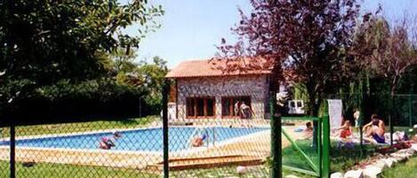 A swimming pool area with a fenced deck, a small building with a red roof, and people relaxing around the pool.
