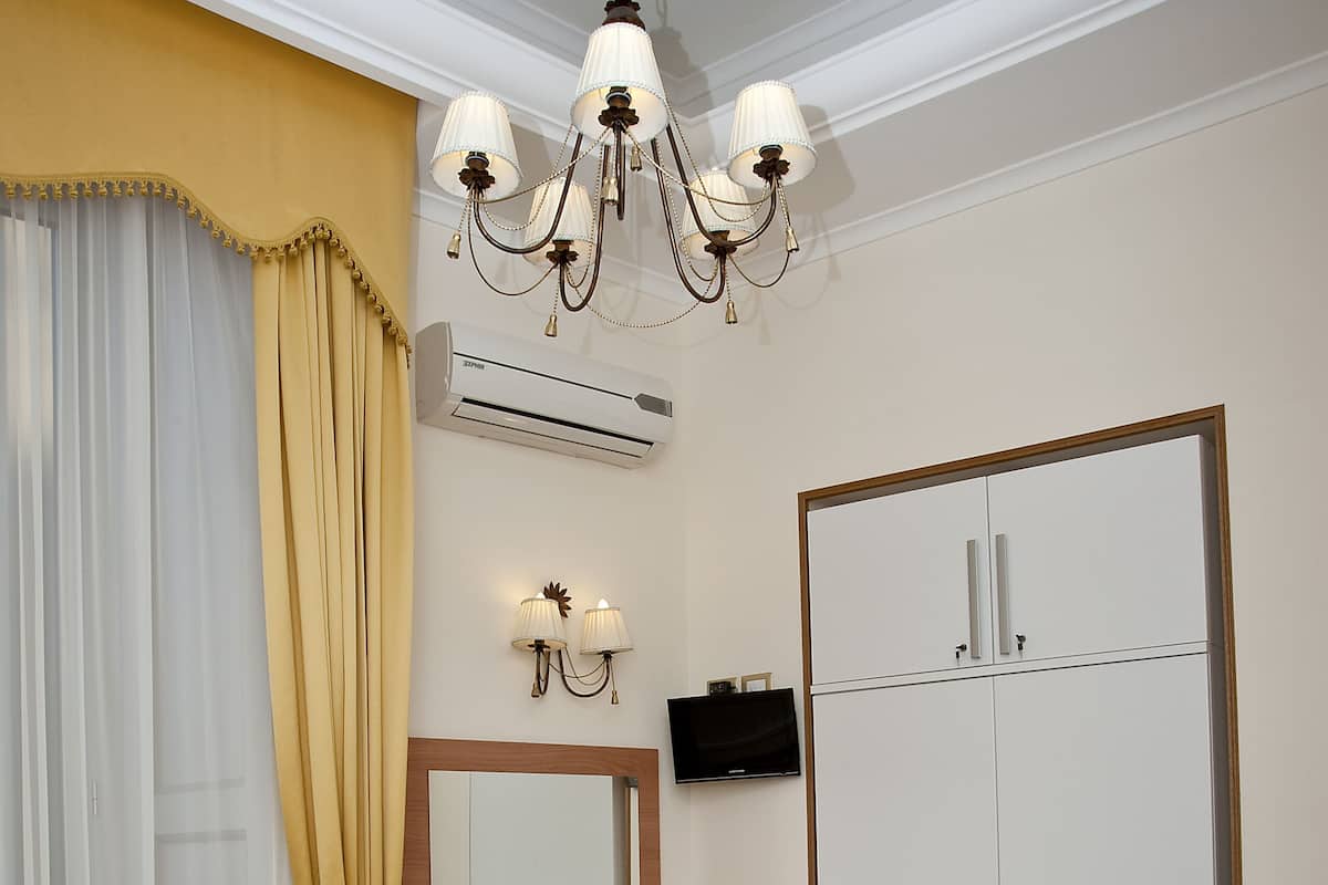 A hotel room with a bed, a desk, a chair, a wardrobe, a chandelier, and an air conditioning unit.