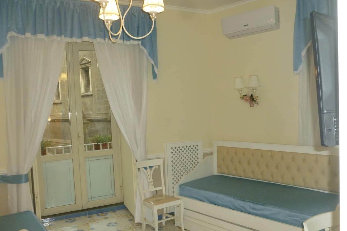 A bedroom with a bed, a small chair, a window with curtains, an air conditioning unit, and a wall-mounted light fixture.