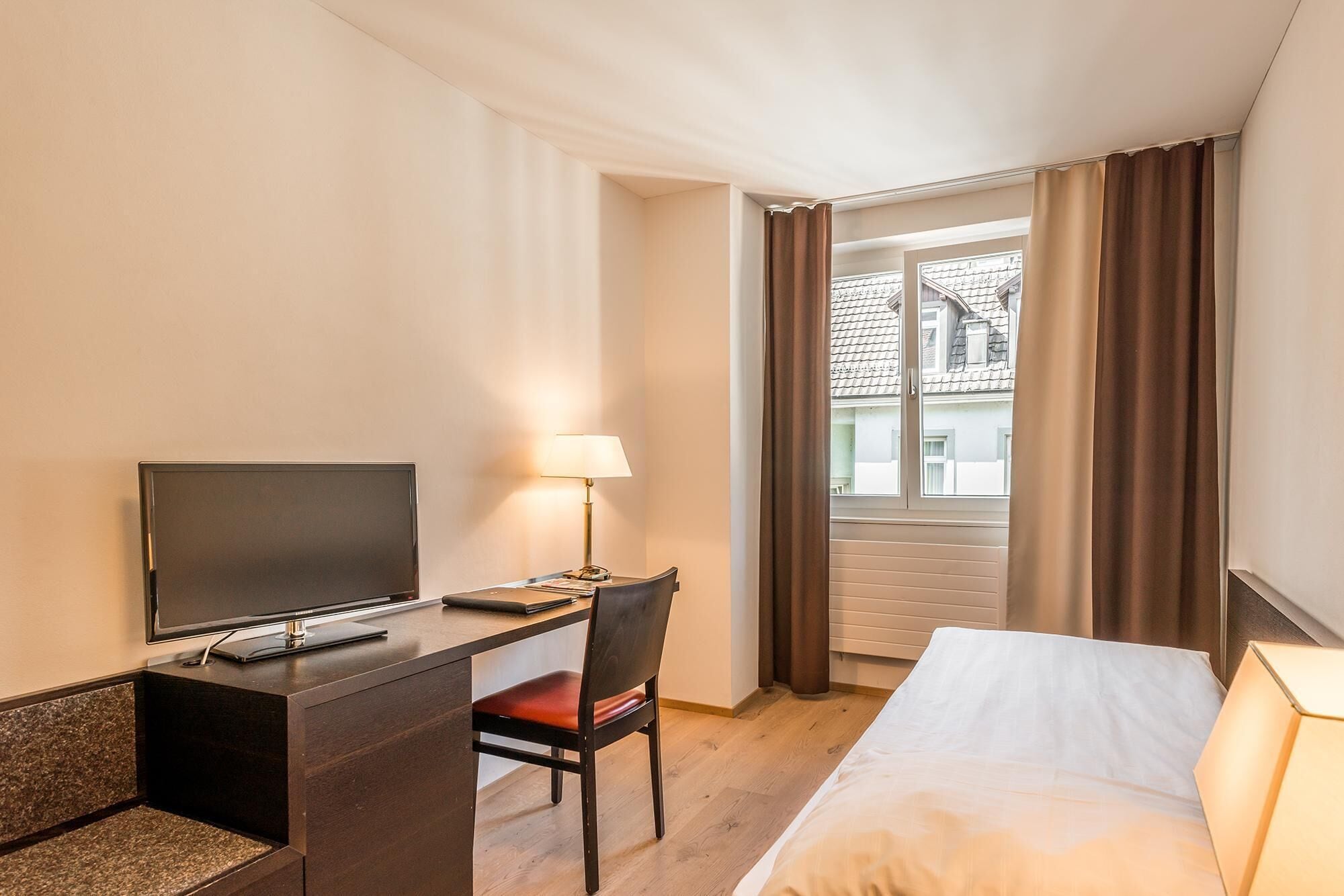 A hotel room with a bed, a desk with a TV, a chair, and a window with curtains.