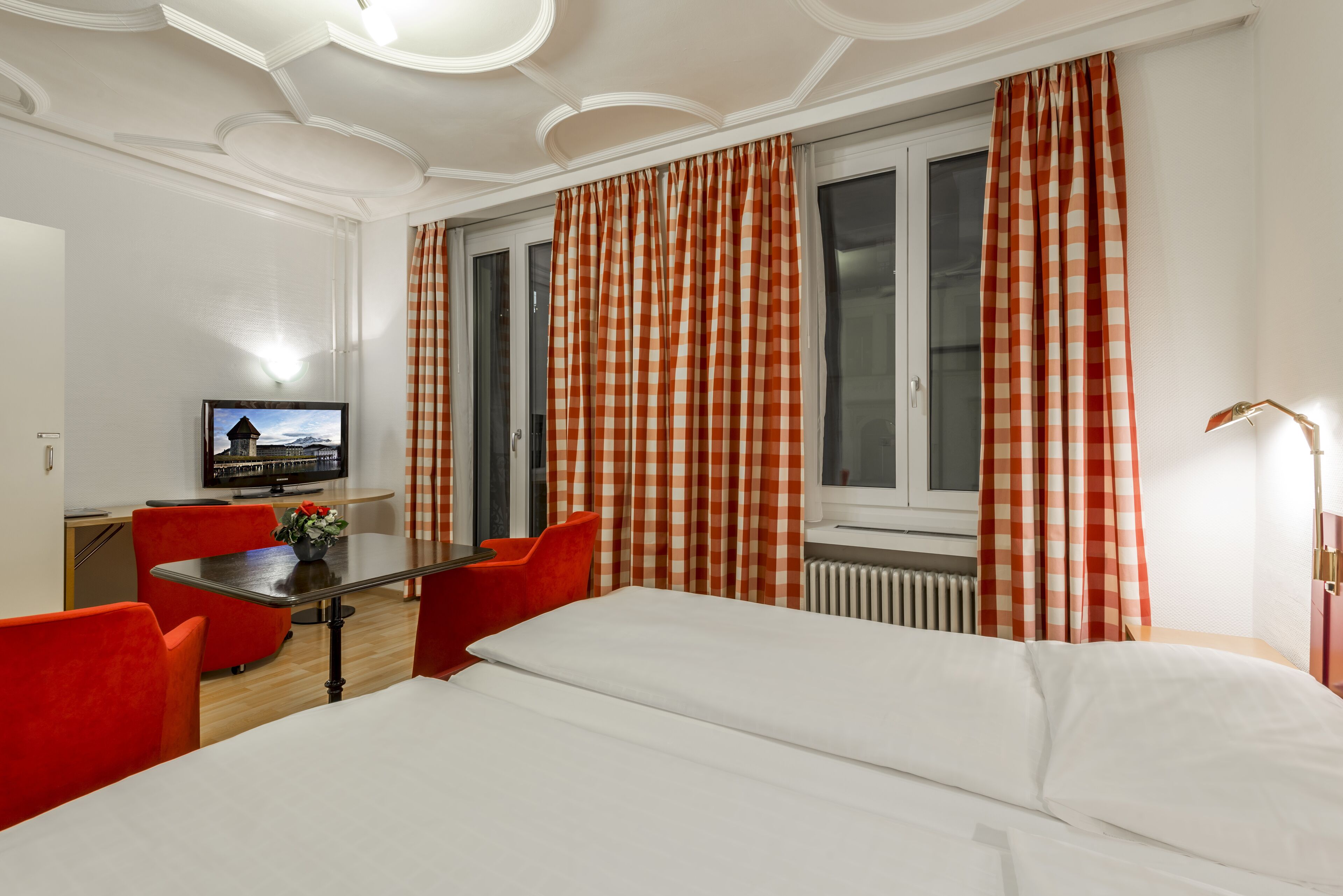 A hotel room with a bed, a desk with a chair, a television, and red chairs.