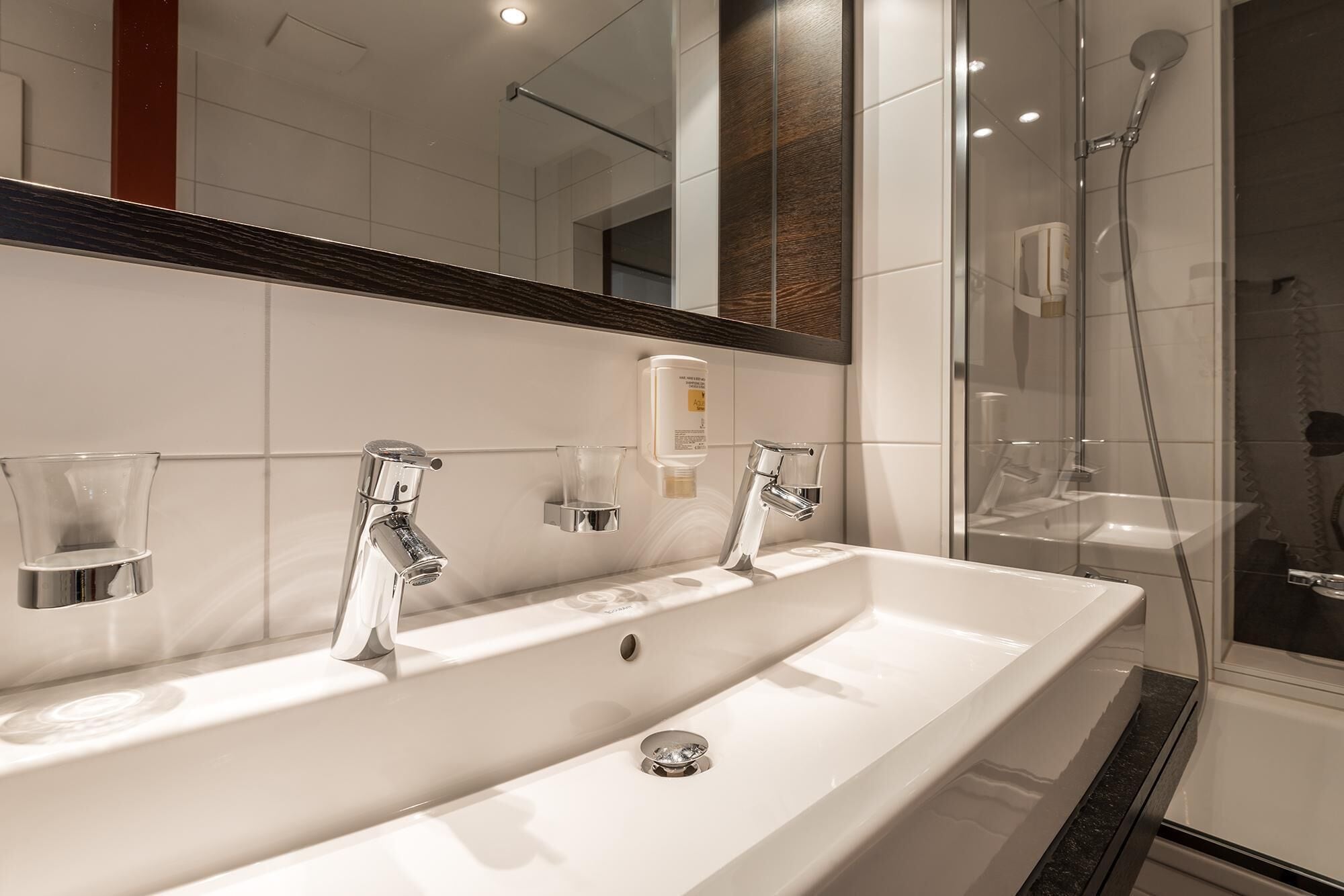 Une salle de bain moderne avec un grand lavabo blanc, deux robinets chromés et une douche séparée par une paroi de verre.