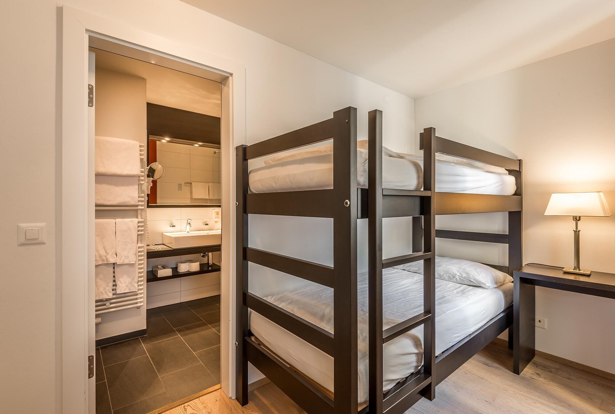 A bunk bed with a bedside table and a lamp, a bathroom with a sink and mirror, and a closet with hanging towels.