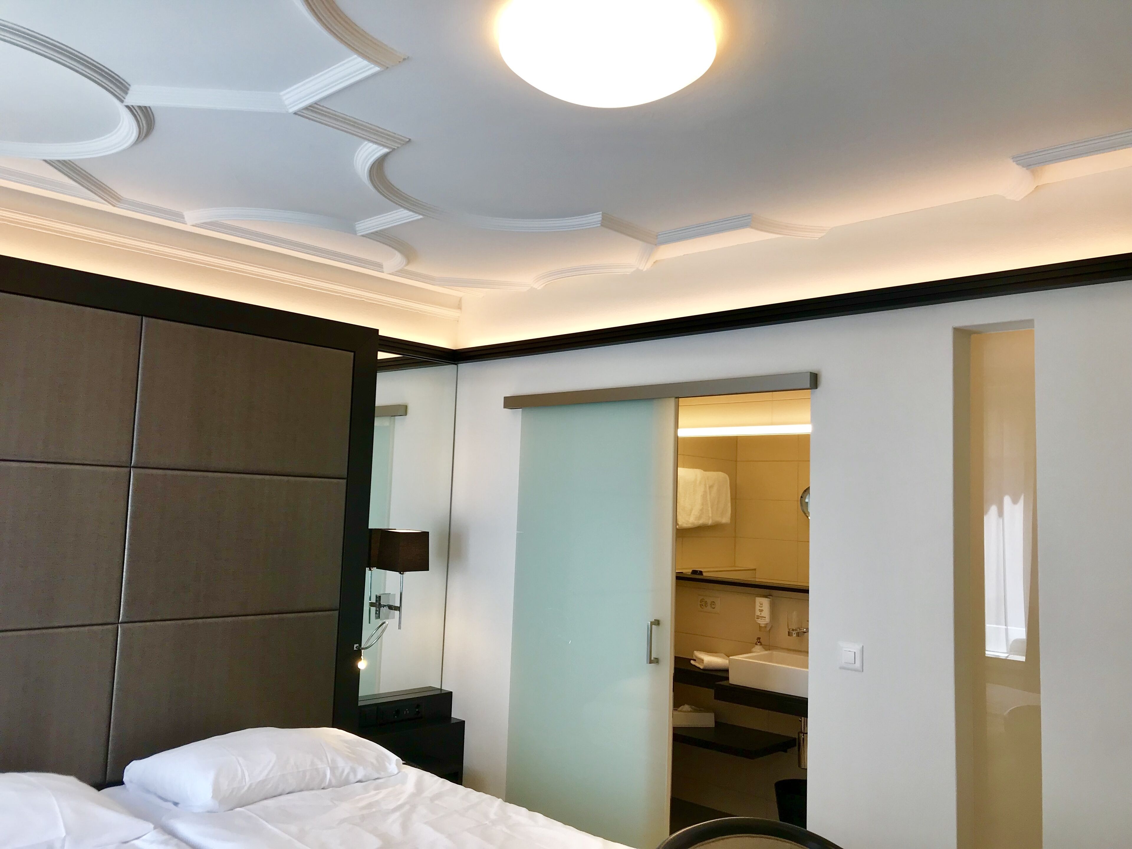 Une chambre d’hôtel moderne dotée d’un grand lit, d’une armoire, d’une salle de bain avec un lavabo et un miroir, et d’un plafond équipé d’un système d’éclairage encastré.