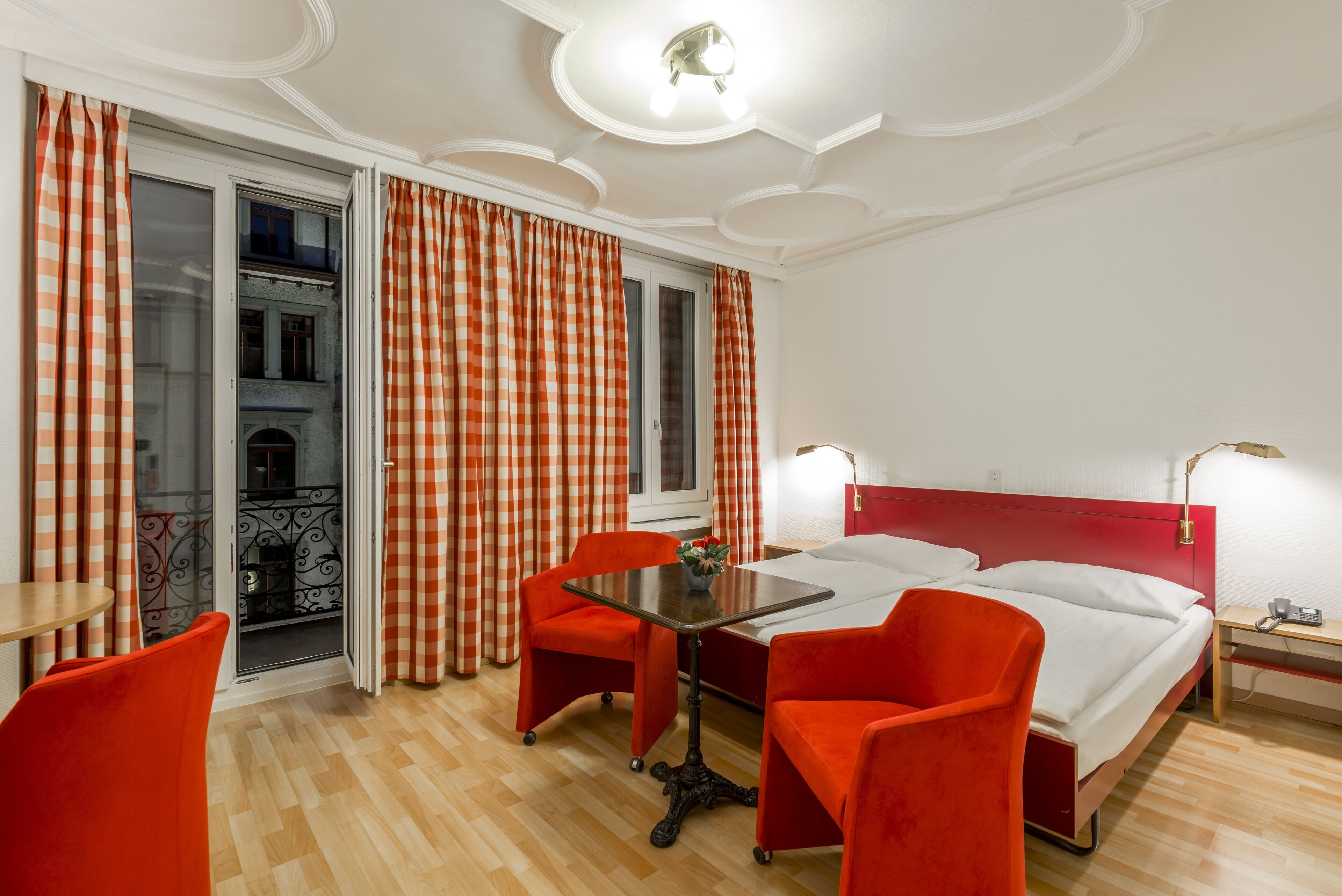 Ein Hotelzimmer mit Bett, zwei Sessel, einem Tisch, einem Balkon mit karierten Vorhängen und einem roten Kopfteil.