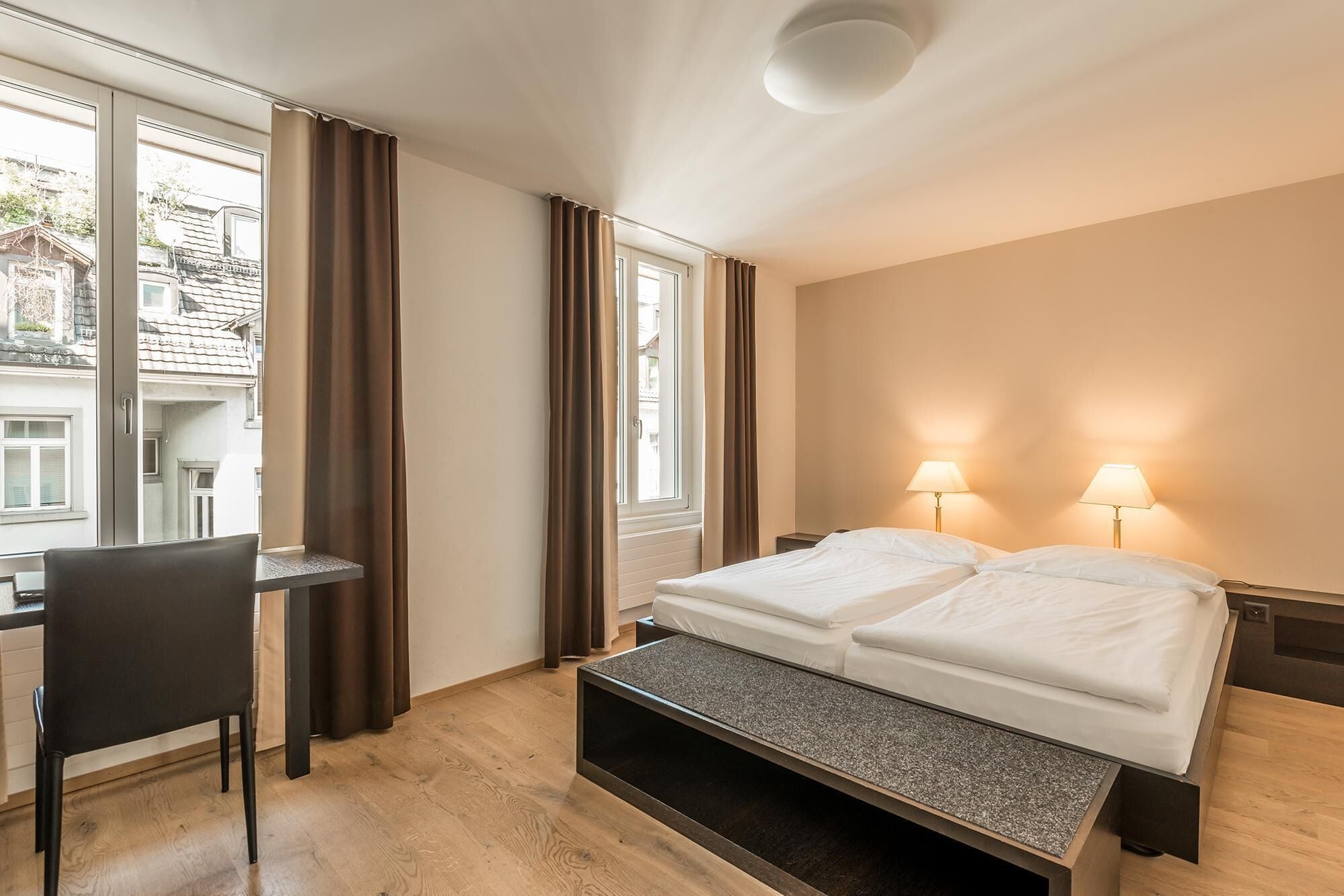 A hotel room with a bed, bedside lamps, a desk, and a chair.