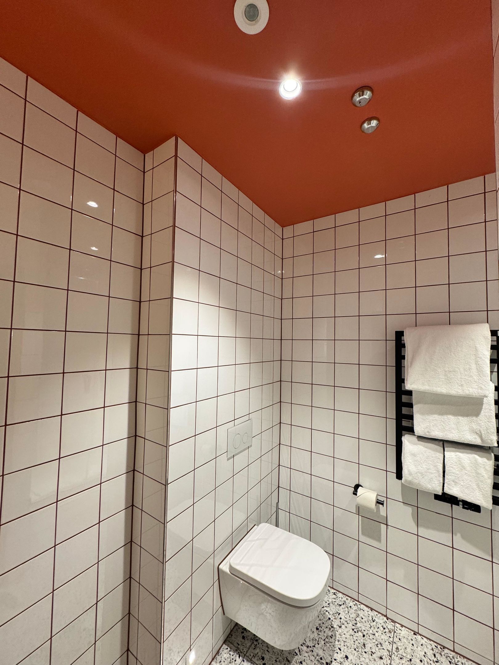 Un baño con paredes de azulejos blancos, un inodoro blanco, un perchero con toallas y una lámpara en el techo.