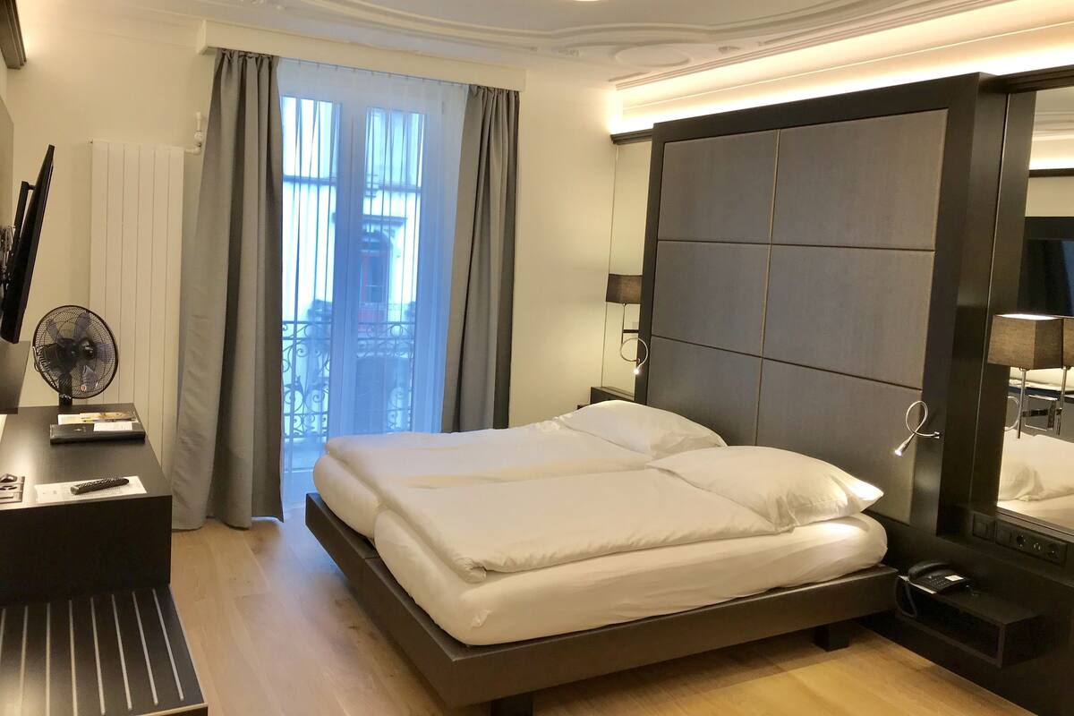 A hotel room with a large bed, a bedside table with a lamp, a mirror, and a view of the outside through a window with curtains.