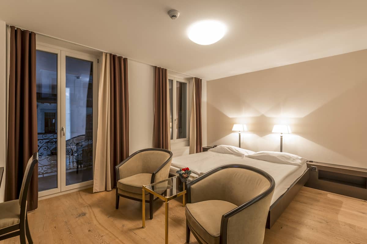 A hotel room with a bed, two chairs, a small table with a vase, and a balcony with curtains.