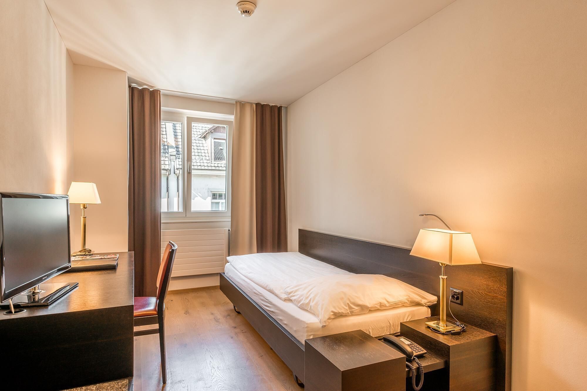 Habitación de hotel con cama, escritorio con televisor, silla, mesita de noche con lámpara y ventana con cortinas.