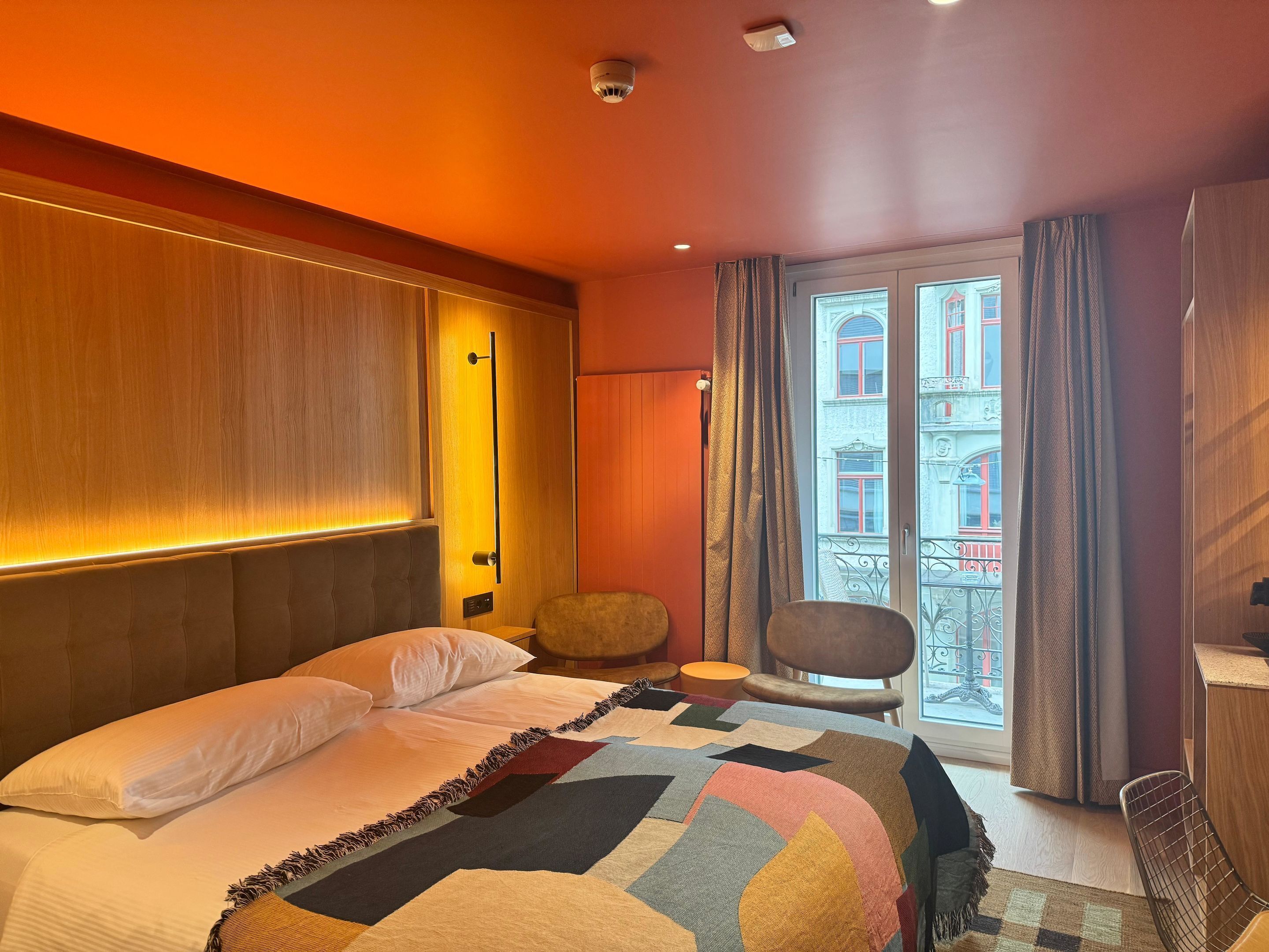A hotel room with a bed, a chair, a window with a view of buildings, and a door with a handle.