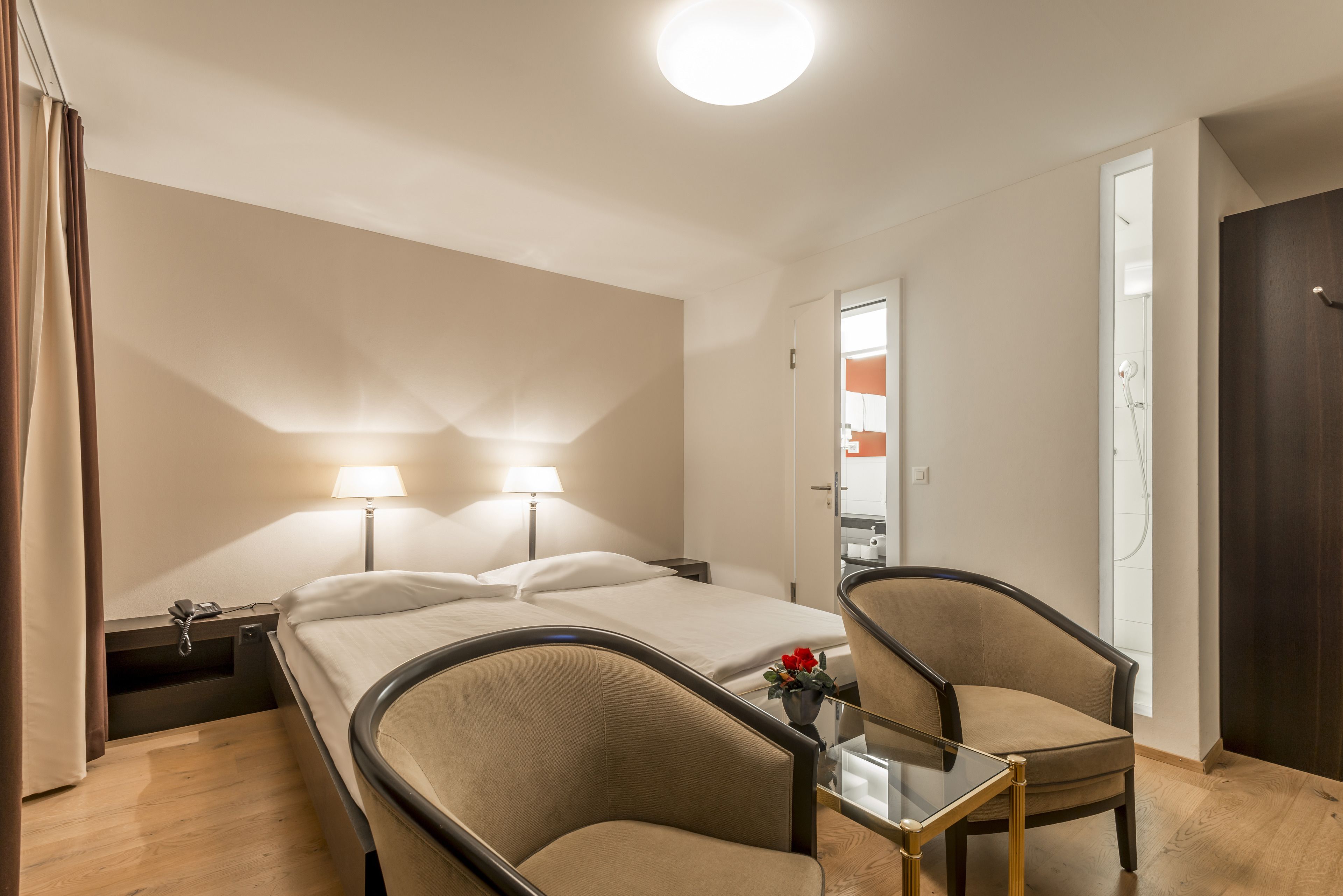 A hotel room with a bed, two bedside lamps, a chair, a small table, and a bathroom visible through an open door.