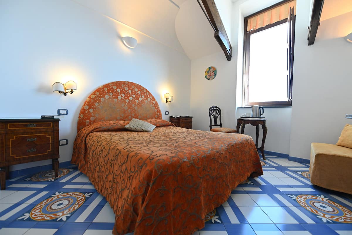 Ein Schlafzimmer mit einem großen Bett, einem Nachttisch, einem Sessel, einem Fenster mit Vorhängen und einer Wandlampe.