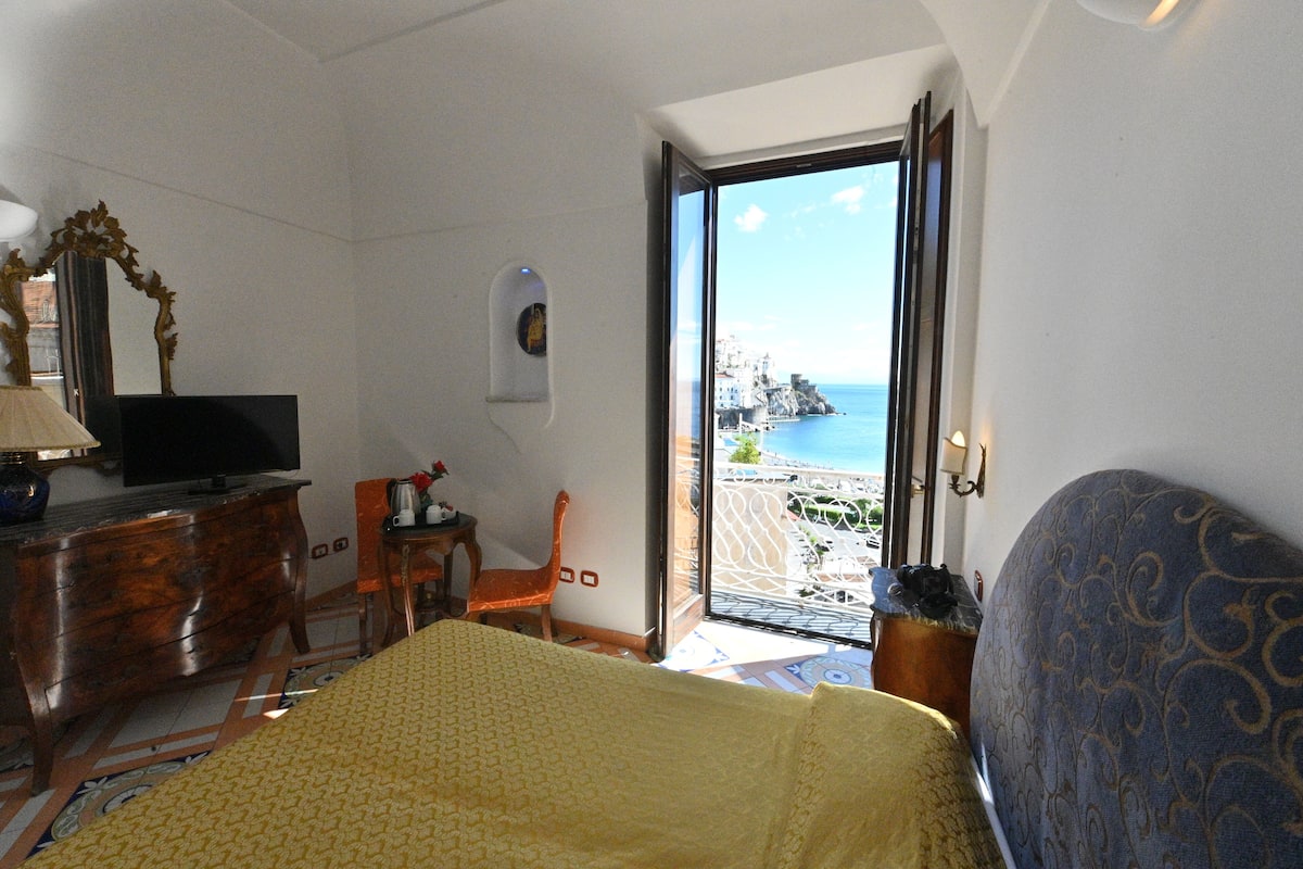 Ein Hotelzimmer mit Bett, Fernseher, Schreibtisch mit Stuhl und einem Balkon mit Meerblick.
