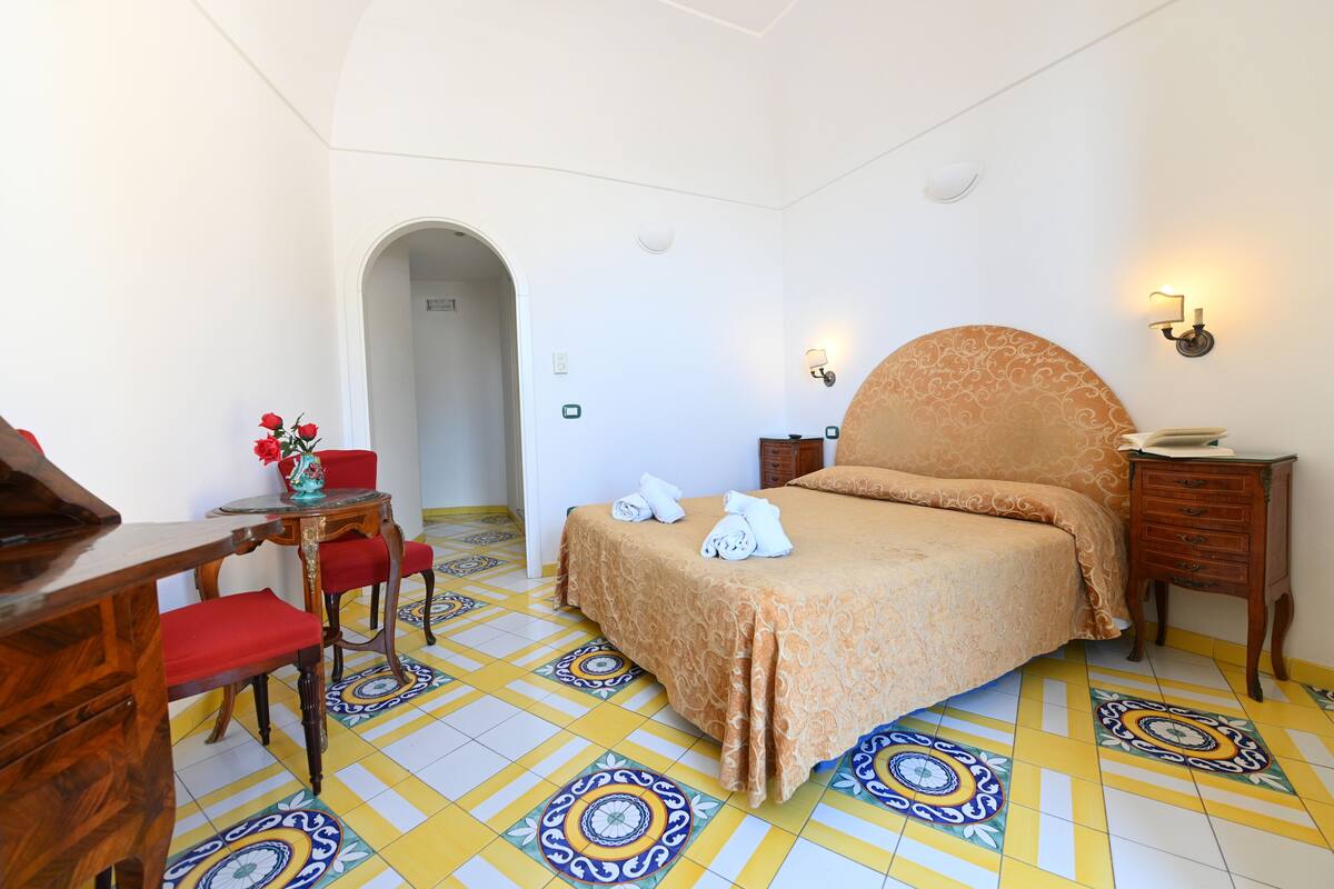 Ein Hotelzimmer mit einem Bett, einem Schreibtisch, einem Stuhl, einem kleinen Tisch mit Blumenvase und einem gemusterten Boden.