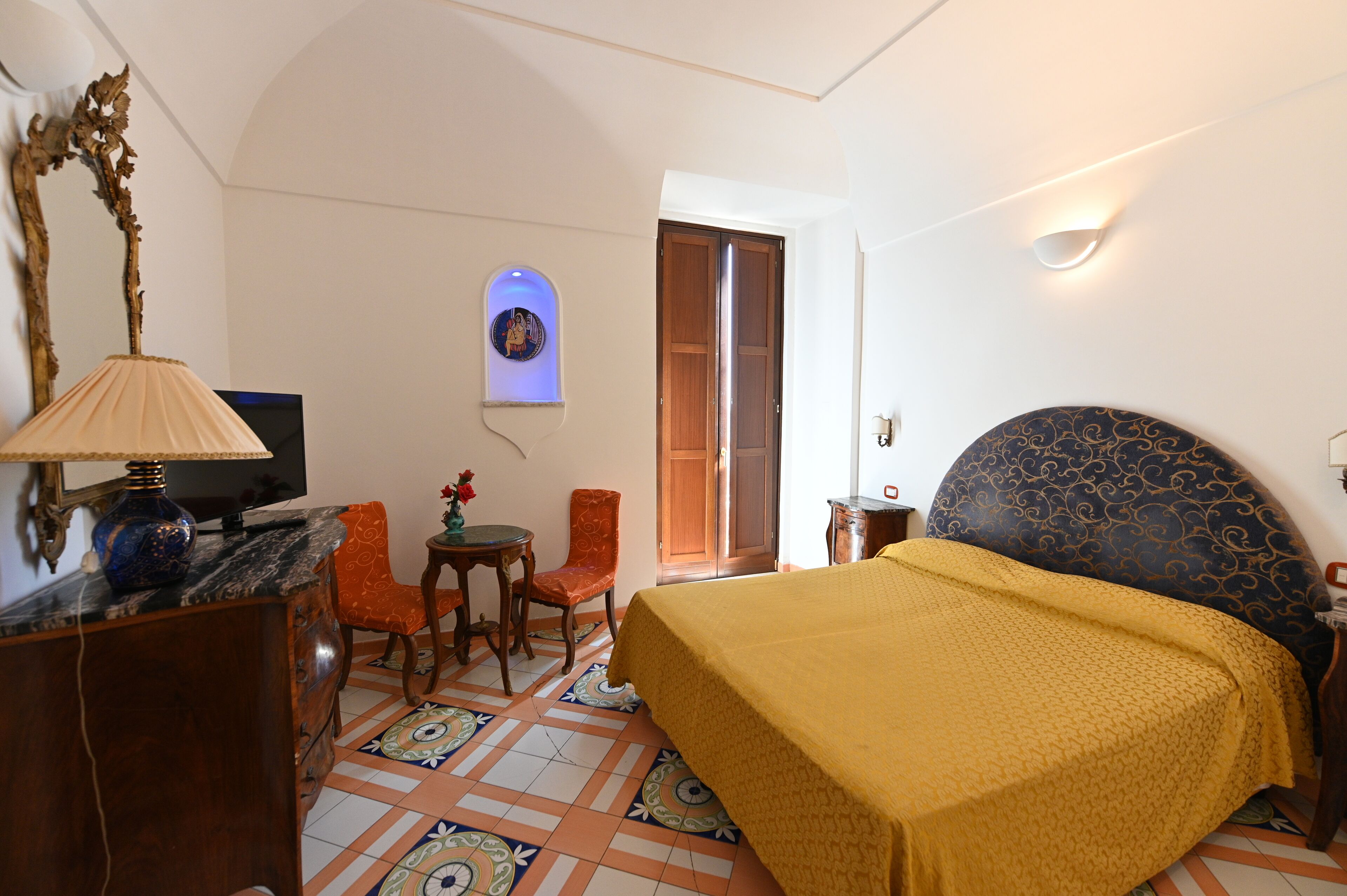 A hotel room with a bed, a desk with a lamp, a chair, and a small table with a vase.