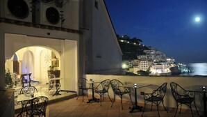 A terrace with outdoor seating, a view of a coastal town at night, and a moon in the sky. - Hotel Croce di Amalfi (Amalfi)