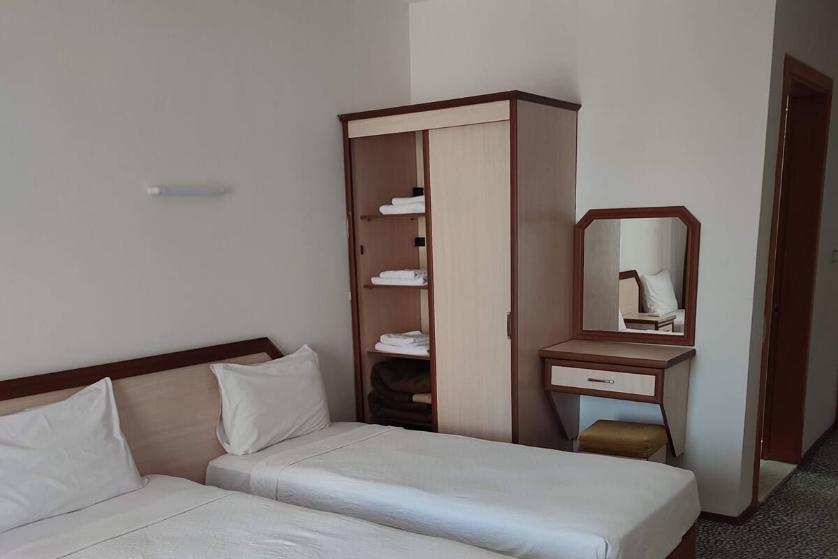 Hotellihuone, jossa on kaksi sänkyä, vaatekaappi, kirjoituspöytä peilillä sekä pieni jakkara.