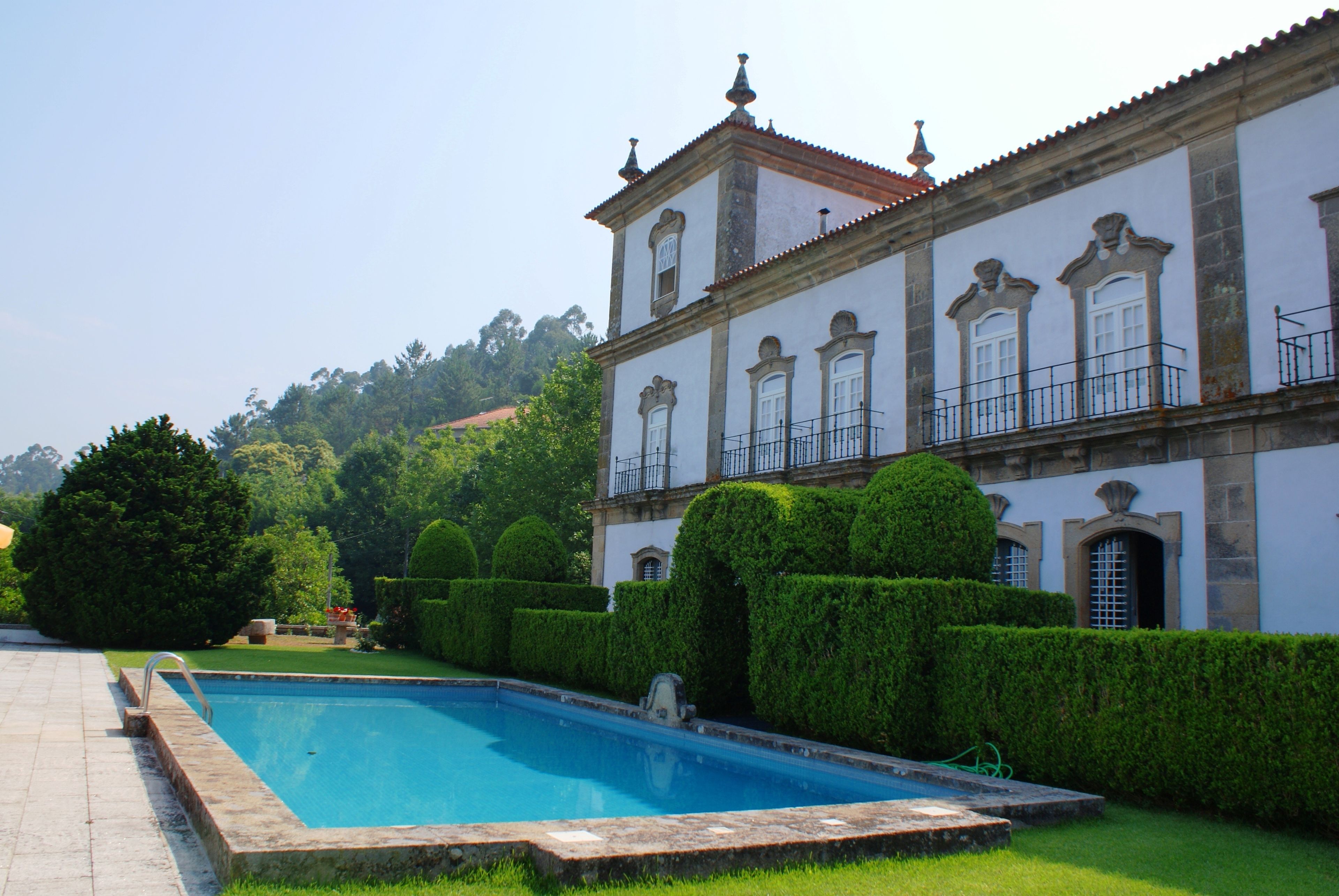 A swimming pool with a stone border, surrounded by well-manicured hedges and a building with classical architecture.