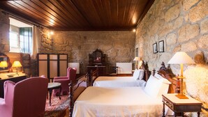 Una habitación acogedora con pared de piedra, techo de madera y una cama con sábanas blancas. Hay dos sillones y una mesita con lámparas.