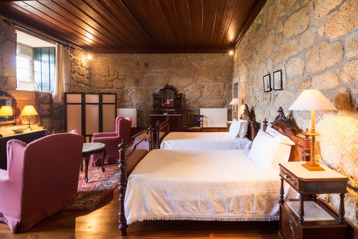 A cozy room with a stone wall, wooden ceiling, and a bed with white linens. There are two chairs and a small table with lamps.