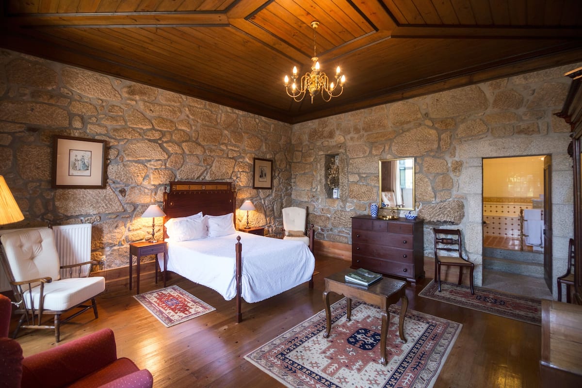 A bedroom with a stone wall, wooden ceiling, a bed with white linens, a wooden dresser, and a chandelier.