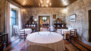 Un comedor rústico con paredes de piedra, techo de madera y una araña. Las mesas están dispuestas para comer, y hay sillas alrededor.