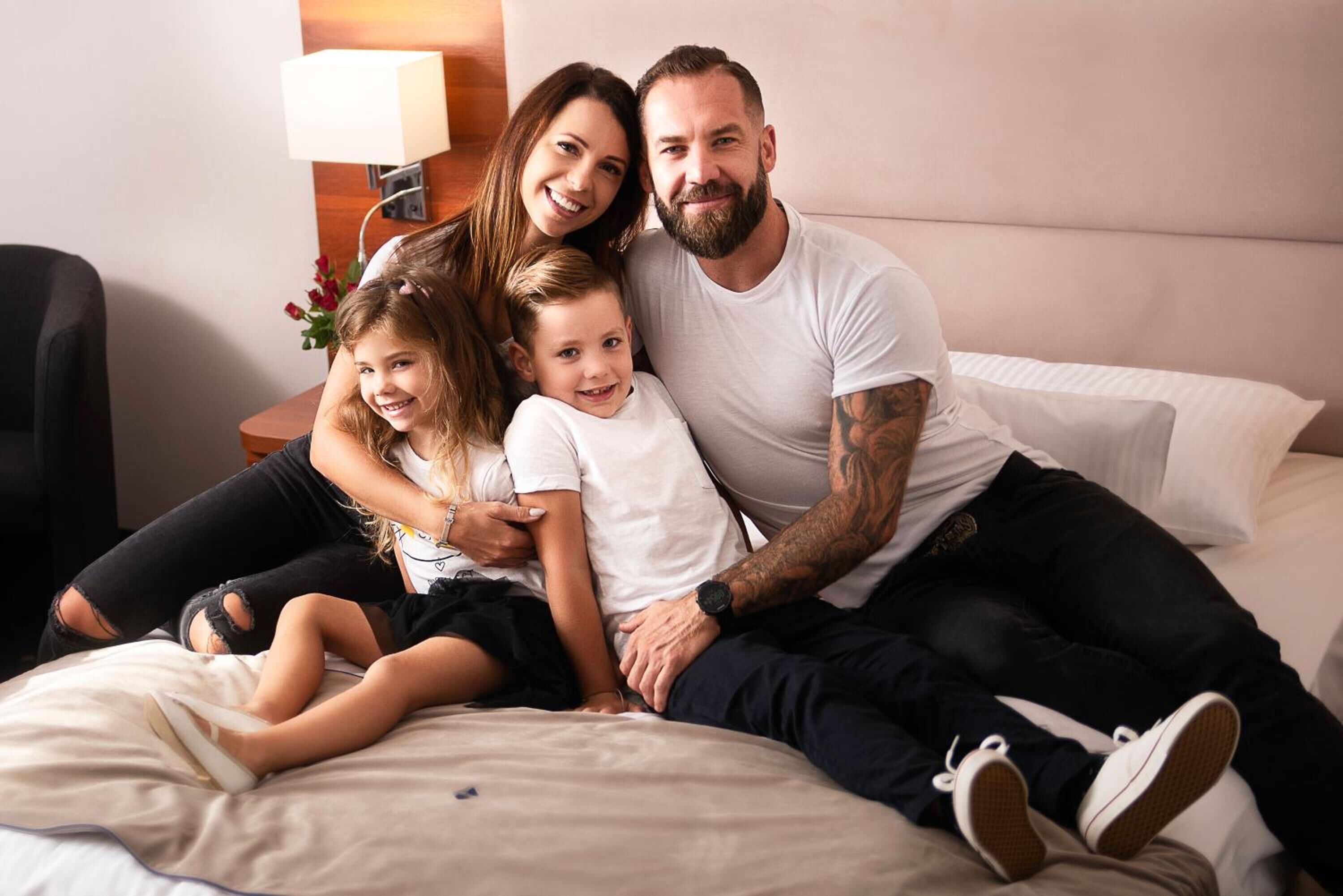 A family of four on a bed in a hotel room with a bedside table and lamp.